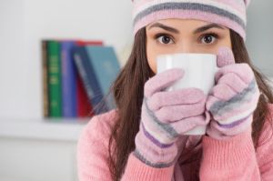Woman holding a mug and bundled up in a cap and gloves in the house